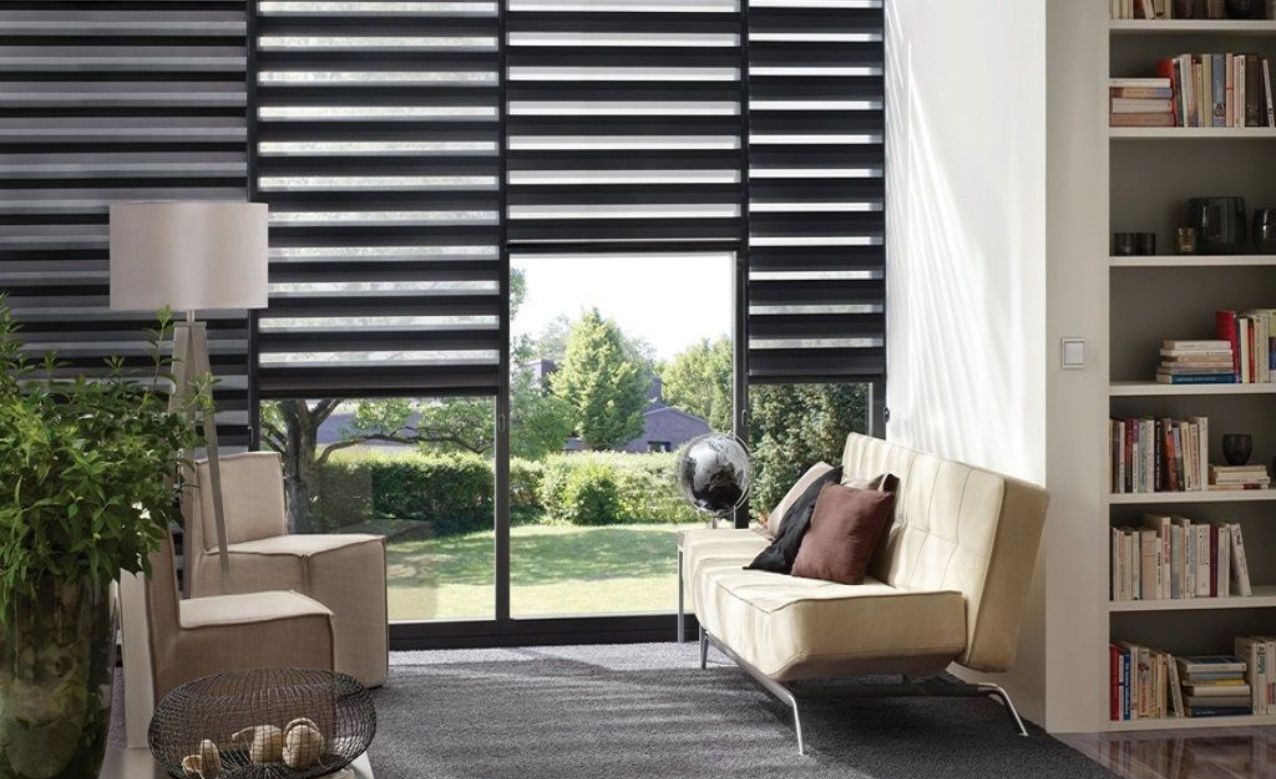 Modern living room with large windows, black horizontal blinds, beige sofas, and a view of a green garden.