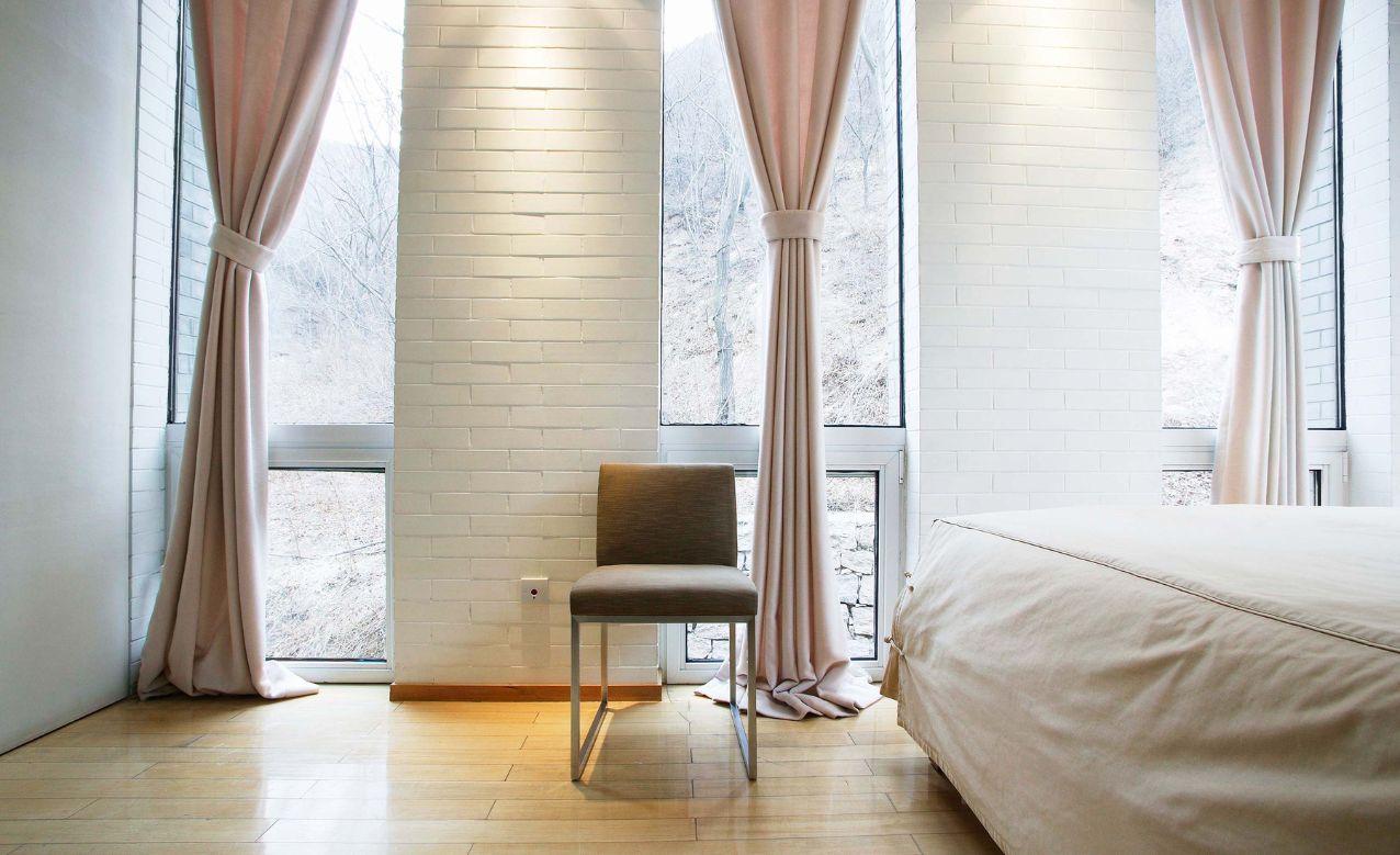 Minimalist bedroom with large windows, pink curtains, a chair, and a light beige bed.